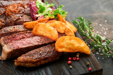 Steaks. Sliced grilled meat steak New York, Ribeye or Chuck roll with with garnished with salad and french fries on black marble board on old wooden background. Top view. Mock up.