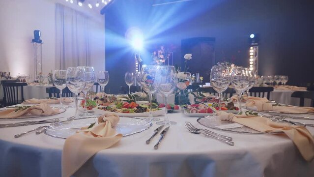 Set festive table with empty glasses, plates and composition of fresh flowers and pastel colored textile napkins. Banquet hall illuminated by rays of rotating spotlight in haze.