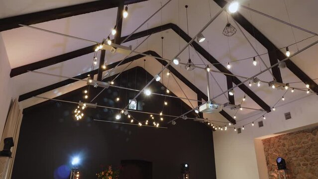 Ceiling of spacious banquet hall with beams, lighting, lanterns and stretched garland. Combined truss system made of wood and metal. Spotlights rotate and illuminate venue with stone and black wall.