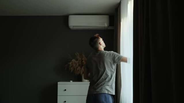 Man closes window in apartment room, takes remote control and remotely turns on air conditioner. Climate device opens blades, blinds, blowing cool air on dry plant in vase on the chest of drawers.