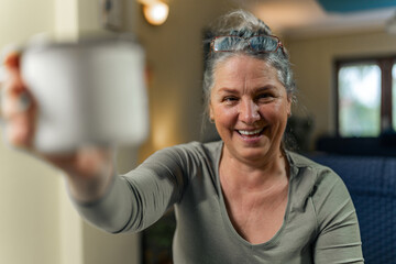 Happy beautiful mature older woman grey-haired drinking coffee, smiling and holding a coffee cup to the camera