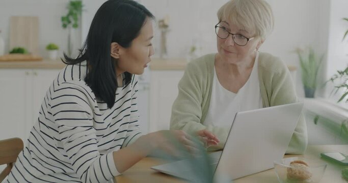 Young daughter teaching senior mother how to use computer talking and typing in kitchen at home. Modern technology and old age concept.