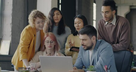 Diverse business team standing around laptop in open space office talking laughing brainstorming. Teamwork and modern technology concept.