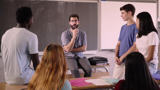 Confident teacher with beard and glasses explaining lesson to runids students at school. Young male trainee chats with his students about the subject matter. University people in tutoring class.