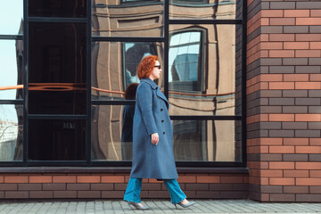 Attractive red-haired woman in a blue coat, jeans and a loose shirt walking along a brick building...