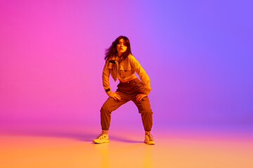 Fototapeta na wymiar Active, artistic young woman, hip hop dancer in sport style clothes dancing against gradient pink purple background in neon light. Concept of contemporary dance, youth, hobby, action and motion