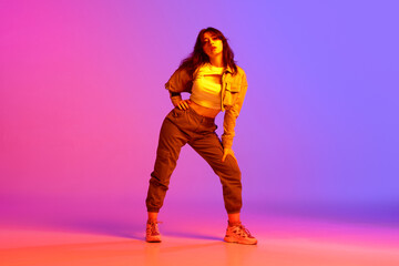 Artistic, beautiful, young girl dancing hip-hop against gradient pink purple background in neon...