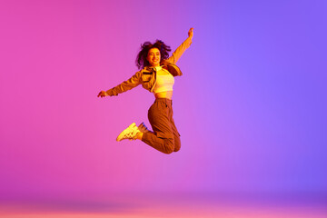 Young girl, hip-hop dancer in sport style clothes jumping against gradient pink purple background in neon light. Delightful. Concept of contemporary dance, youth, hobby, action and motion