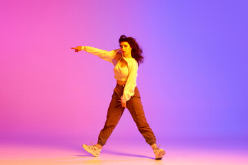 Fototapeta na wymiar Dynamic image of young sportive girl, female hip-hop dancer training against gradient pink purple background in neon light. Concept of contemporary dance, youth, hobby, action and motion