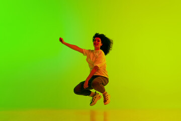 Fototapeta na wymiar Young girl, hip-hop dancer in sport style clothes jumping against gradient green yellow background in neon light. Concept of contemporary dance, youth, hobby, action and motion