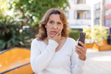 middle age pretty woman with mouth and eyes wide open and hand on chin. using her smartphone