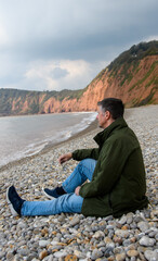 Fototapeta premium A man sits on a beach in front of a cliff, staring out to sea. Concept image, he ponders his retirement, finances, health and pensions. He is isolated and alone. portrait view, copy space. Editorial.