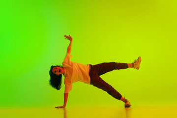 Fototapeta na wymiar Artistic, flexible, talented young girl, hip-hop dancer performing against gradient green yellow background in neon light. Concept of contemporary dance, youth, hobby, action and motion