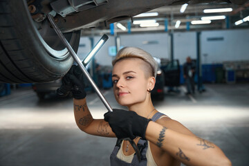 Fototapeta na wymiar Pretty woman in work overalls is repairing car on lift