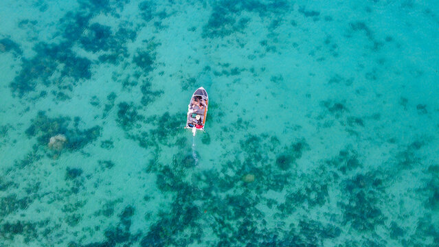 Drone shot of small tinny with people driving through turquoise waters with sand and coral below
