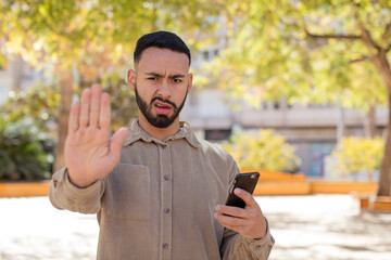young  adult man looking serious showing open palm making stop gesture. using a smartphone concept