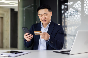 Fototapeta na wymiar Successful asian boss working inside office with laptop at workplace, businessman holding phone and bank credit card, man using app for booking and online purchase money transfer.