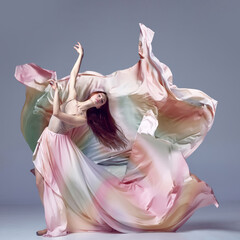 Inspired young, beautiful ballerina wearing rainbow dress dancing over grey studio background. Ballet with silk dress