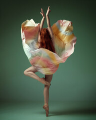 Young classical dancer wearing colorful flying dress dancing on fingertips over dark green studio background. Tender movement © Lustre Art Group 