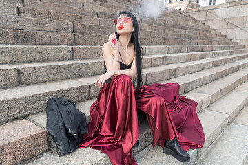 A sexy woman and a bright red satin skirt is sitting on the steps. leather jacket nearby. She smokes an electronic cigarette.
