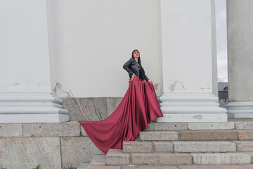 Woman with long hair, perfect figure, sexy. she is in a satin, long red skirt, leather jackets. Posing in the city at the tall ancient columns