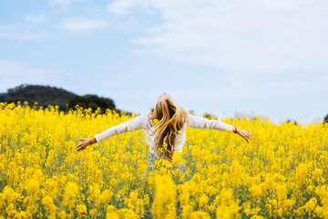 Rear view of a blonde young woman looking at the sky with arms open amidst a field of blooming yellow rapeseed flowers