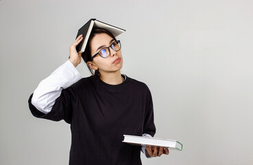 Young pretty student girl with glasses holding book on her head. She holds a stack of books for reading, studying or education. High quality isolated photo on gray background.