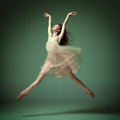 Young and incredibly beautiful ballerina wearing tulle dress jumping gracefully over dark green studio background. Demonstrate flexibility - obrazy, fototapety, plakaty