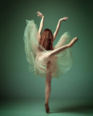 Back view of one adorable ballet dancer, woman wearing elegant dress dancing on tiptoes over dark green studio background