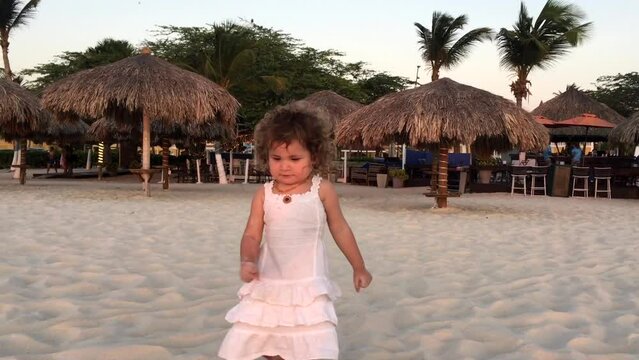 Little girl in white dress walking on sand beach on Aruba island in Caribbean Sea. Beautiful sunset light. Palm trees and traditional beach umbrellas behind. High quality FullHD footage