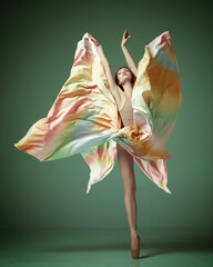 Portrait of young ballerina, ballet dancer wearing colorful dress dancing on tiptoe over dark green studio background. Aesthetic of movement