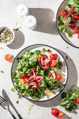 Green salad with ham or prosciutto, corn salad leaves, tomatoes and cheese in hard light with clear shadows. Healthy diet lunch on white marble background top view flat lay