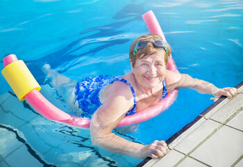 Active senior (elderly) woman (over age of 50) in sport goggles, swimsuit doing aqua fitness with swim noodles in swimming pool. Mature female smiling in summer day. Healthy lifestyle.