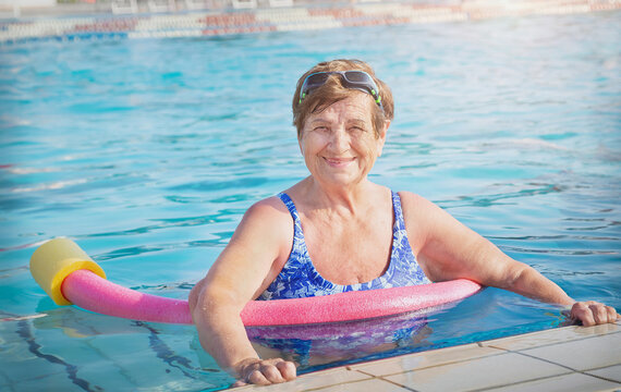Active senior (elderly) woman (over age of 50) in sport goggles, swimsuit and with swim noodles in swimming pool before water aerobics. Healthy lifestyle.