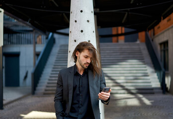 Fototapeta premium Modern business concept using mobile devices. Attractive young business man in black suit using smartphone on the street, ready to tackle any task at any time.