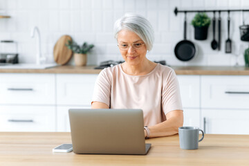 Positive pretty modern caucasian middle aged gray haired woman with glasses, sits at home in the kitchen, uses laptop for remote work, online shopping, read news, looks satisfied, smiles