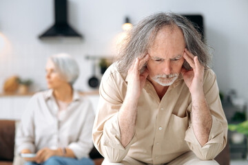 Mature caucasian spouses misunderstanding each other, experiencing crisis in relationship, offended husband looks away disappointedly, the wife sits in defocus on a background. Quarrel, marital crisis