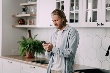 handsome blond man stands in the kitchen and uses a smartphone