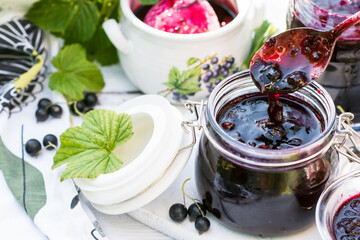 Cooked homemade black currant jam on spoon above jar and white wooden table and vivid tablecloth background, fresh black currant jam close up, food and healthy eating concept