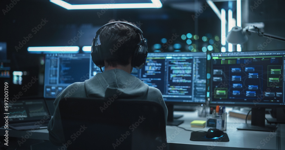 Poster System Administrator Working in a Dark Research Facility on a Computer with Multiple Displays. Software Developer in Casual Clothes Wearing Headphones and Updating Server System Database