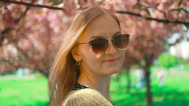 Cute blonde woman is enjoying a spring blossom. Happy smiling girl looking at the camera. A sunny spring in the fresh air. Spring blooms of a sakura tree in a city park. Super slow motion photography.