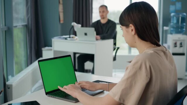 Young Woman Uses Laptop with Green Mock-up Screen In Office. Business Lady or IT Specialist Works Sitting at Table with Notebook