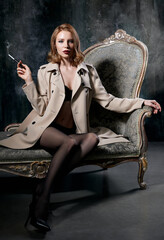 Fototapeta na wymiar Film noir style: dangerous elegant young woman sitting on sofa and smoking cigarette. Vintage (retro) portrait of sexy lady in black stockings, lingerie and coat