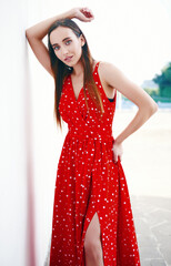 Street fashion: beautiful sweet girl in red dress. Portrait of alluring young woman standing at wall