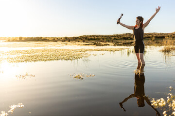 Woman taking selfie with camera in nature in middle of lake, surrounded by flowers, with selective focus, sunset in middle of lake woman traveling alone, waves in water, movie colours.