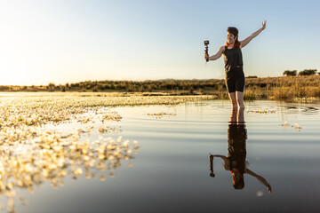 Woman taking selfie with camera in nature in middle of lake, surrounded by flowers, with selective focus, sunset in middle of lake woman traveling alone, waves in water, movie colours.