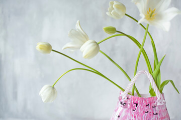 Delicate white flowers tulips in a vase in the shape of a handbag on a gray wall background. Nice interior photo for a poster.