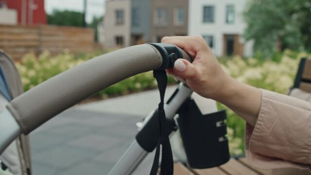 Medium close-up of hand of unrecognizable young mother sitting on bench pulling and pushing baby stroller