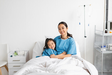 Fototapeta premium cheerful asian mother and child embracing and looking at camera on bed in hospital ward.
