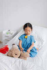 asian child in hospital gown doing injection to teddy bear with toy syringe while playing on bed in clinic.